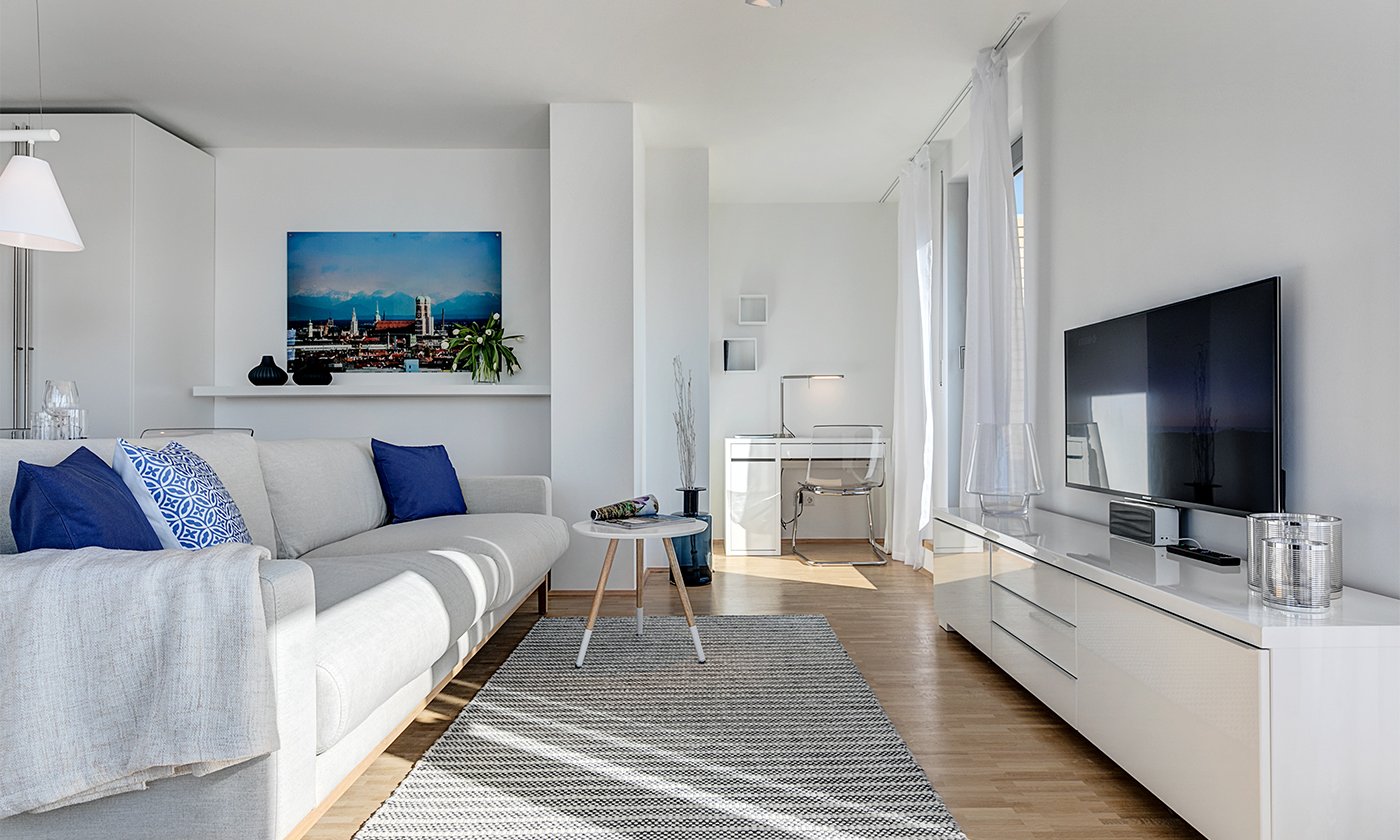 The photo shows a living room with a white sofa in front of a television. A picture of the Munich skyline hangs on the wall.